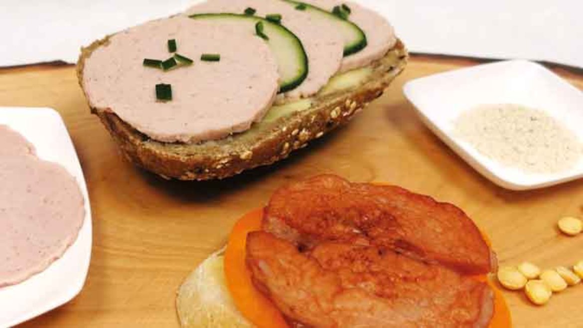 Ein belegtes Roggenbrot mit Scheiben von gekochtem Schinken, Gurkenscheiben und gehackten Kräutern. Daneben liegen zwei Scheiben geröstete Tomate und ein Teller mit Dip sowie einige Nüsse.