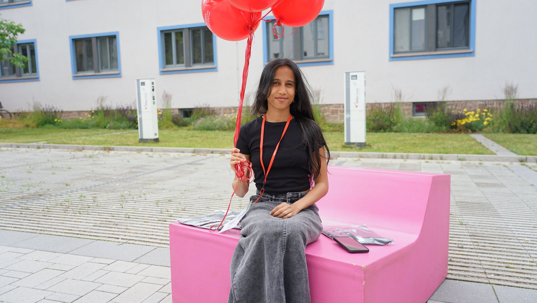 Eine Person sitzt lächelnd auf einer pinken Sitzbank vor einem Gebäude und hält ein Getränk. Sie trägt eine rote Schleife und hält fünf rote Luftballons in der Hand.