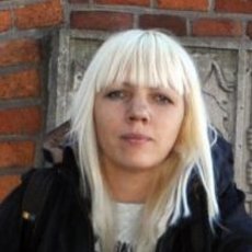 A person with blonde hair styled in a short, blunt cut, wearing a black jacket, standing in front of a brick wall. The person has a neutral expression and is looking directly at the camera.