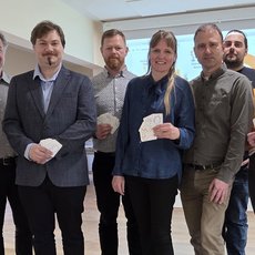 Gruppe von sieben Personen, die jeweils ein Spielkarten-Paket oder eine Spielkarte in der Hand halten und lächelnd in die Kamera blicken.