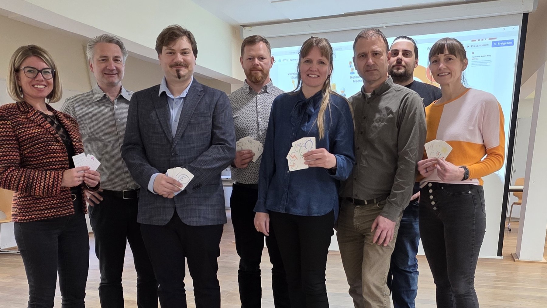 Gruppe von sieben Personen, die jeweils ein Spielkarten-Paket oder eine Spielkarte in der Hand halten und lächelnd in die Kamera blicken.