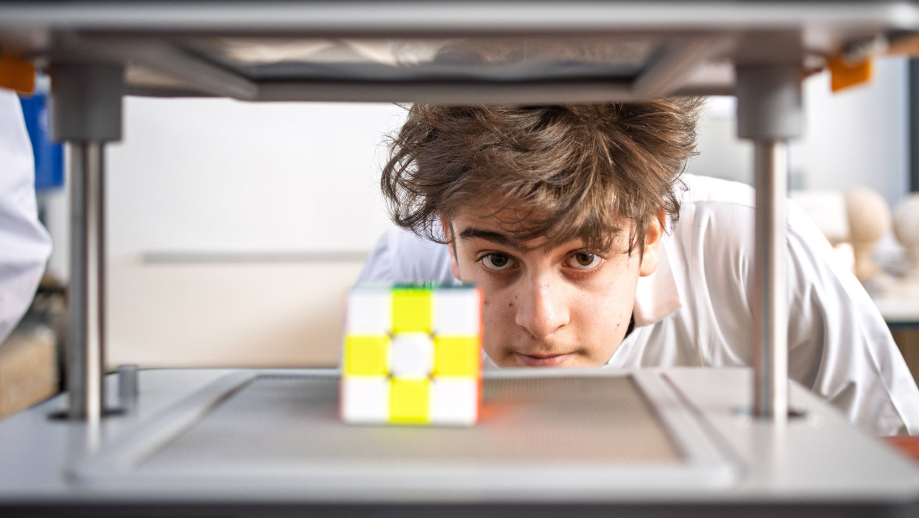 Eine Person in weißem Laborkittel beobachtet konzentriert ein kleines, gelb-weißes Würfelobjekt, das sich in einer transparenten Vorrichtung befindet, vermutlich ein 3D-Drucker oder ähnliche Maschine.