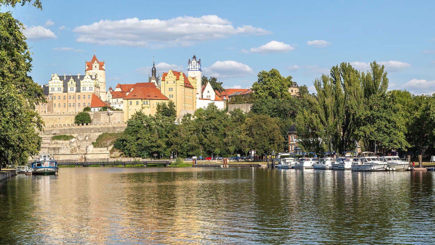 Ein malerisches Bild einer historischen Stadt mit einem Fluss im Vordergrund. Die Stadt zeigt alte Gebäude mit roten Dächern und Türmen, umgeben von grünen Bäumen. Boote sind auf dem ruhigen Wasser zu sehen, und der Himmel ist teilweise bewölkt.