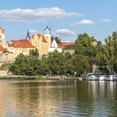 A scenic view of a historic town with a castle-like structure and red-roofed buildings, situated by a calm river. The town is surrounded by lush green trees, and several boats are docked along the riverbank under a partly cloudy sky.