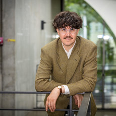 Ein Mann mit kurzem, lockigem Haar, Brille und leichtem Bart lächelt freundlich. Er trägt einen grünen Tweed-Blazer über einem weißen Hemd und sitzt entspannt auf einer Metallgeländer-Stütze vor einem modernen Gebäude mit Glas- und Betonelementen.