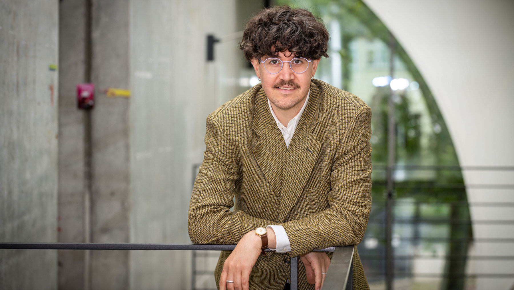 Ein Mann mit kurzem, lockigem Haar, Brille und leichtem Bart lächelt freundlich. Er trägt einen grünen Tweed-Blazer über einem weißen Hemd und sitzt entspannt auf einer Metallgeländer-Stütze vor einem modernen Gebäude mit Glas- und Betonelementen.