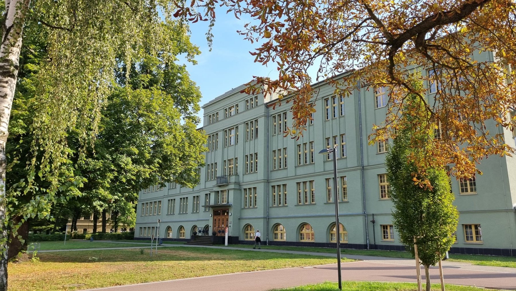 Großes, hellblau gestrichenes Schulgebäude mit klassischer Fassade, zahlreichen Fenstern und zentralem Eingang, umgeben von grünem Rasen und Bäumen mit Herbstfärbung.