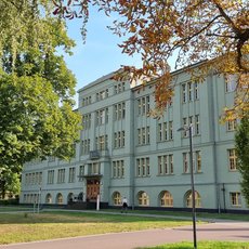 Großes, hellblau gestrichenes Schulgebäude mit klassischer Fassade, zahlreichen Fenstern und zentralem Eingang, umgeben von grünem Rasen und Bäumen mit Herbstfärbung.