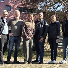 Sechs Personen stehen lässig im Freien vor einem alten Mauergebäude und einem Auto, lächelnd für ein Gruppenfoto. Vier Männer und zwei Frauen, alle in moderner Alltagskleidung.