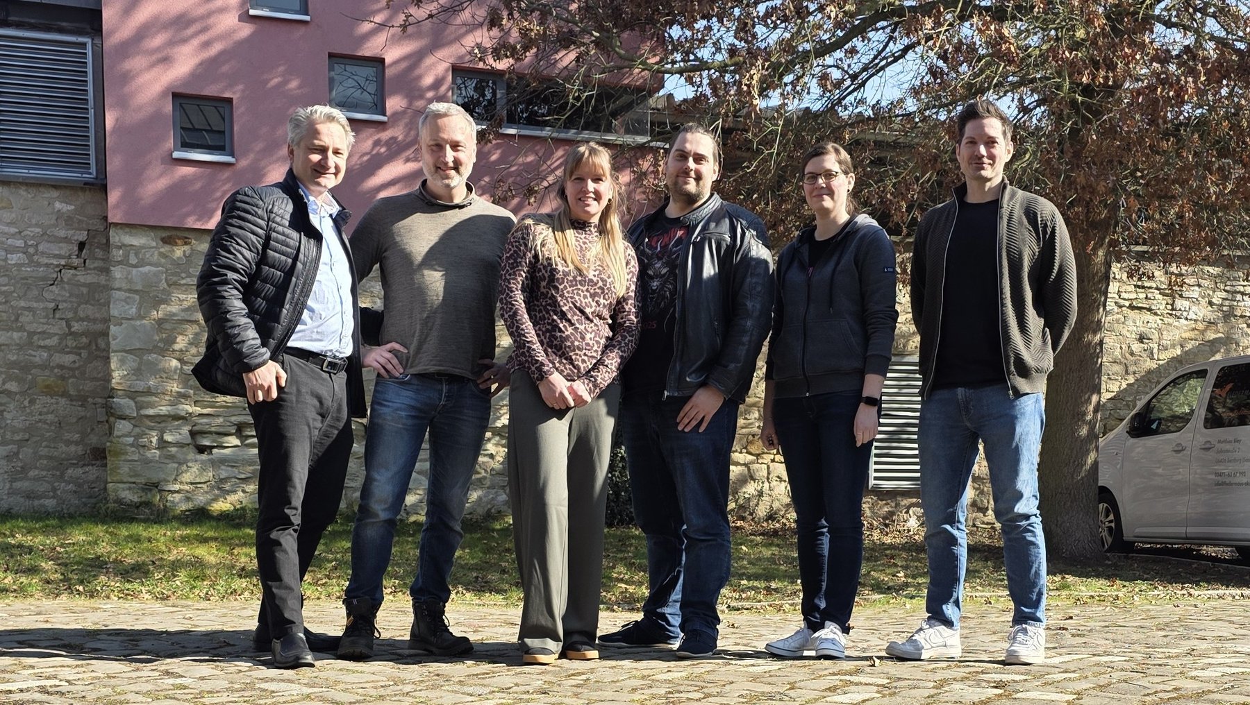 Sechs Personen stehen lässig im Freien vor einem alten Mauergebäude und einem Auto, lächelnd für ein Gruppenfoto. Vier Männer und zwei Frauen, alle in moderner Alltagskleidung.