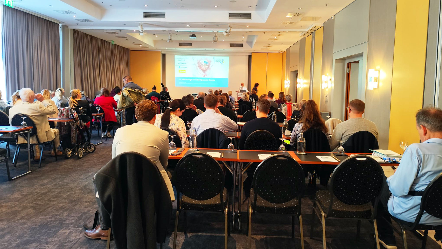Großer Seminarraum mit vielen Teilnehmenden, die an runden Tischen sitzen und sich unterhalten. Im Hintergrund steht eine Person am Podium mit einer Präsentation auf der Leinwand, die ein Diagramm mit blauen und roten Elementen zeigt.