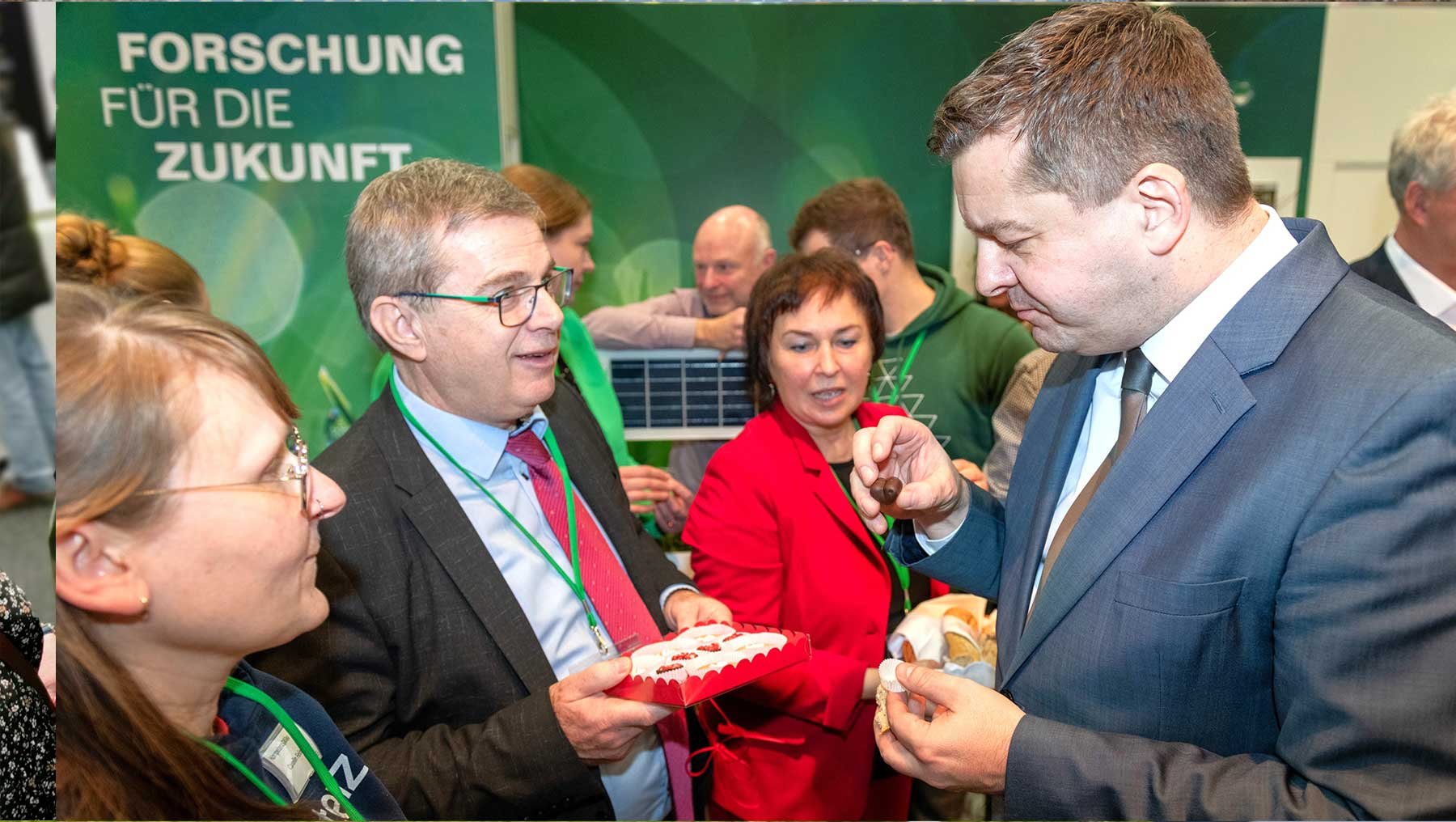 Drei Personen auf einer Veranstaltung halten und probieren kleine, runde Gebäckstücke, während im Hintergrund weitere Teilnehmer und ein Banner mit der Aufschrift „Forschung für die Zukunft“ zu sehen sind.