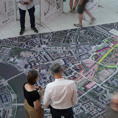 Großflächiger Stadtplan auf dem Boden mit markierten Wegen und Gebäuden, um den mehrere Personen in einer Ausstellung stehen und sich unterhalten. Im Hintergrund hängen weitere Stadtpläne und Infotafeln.