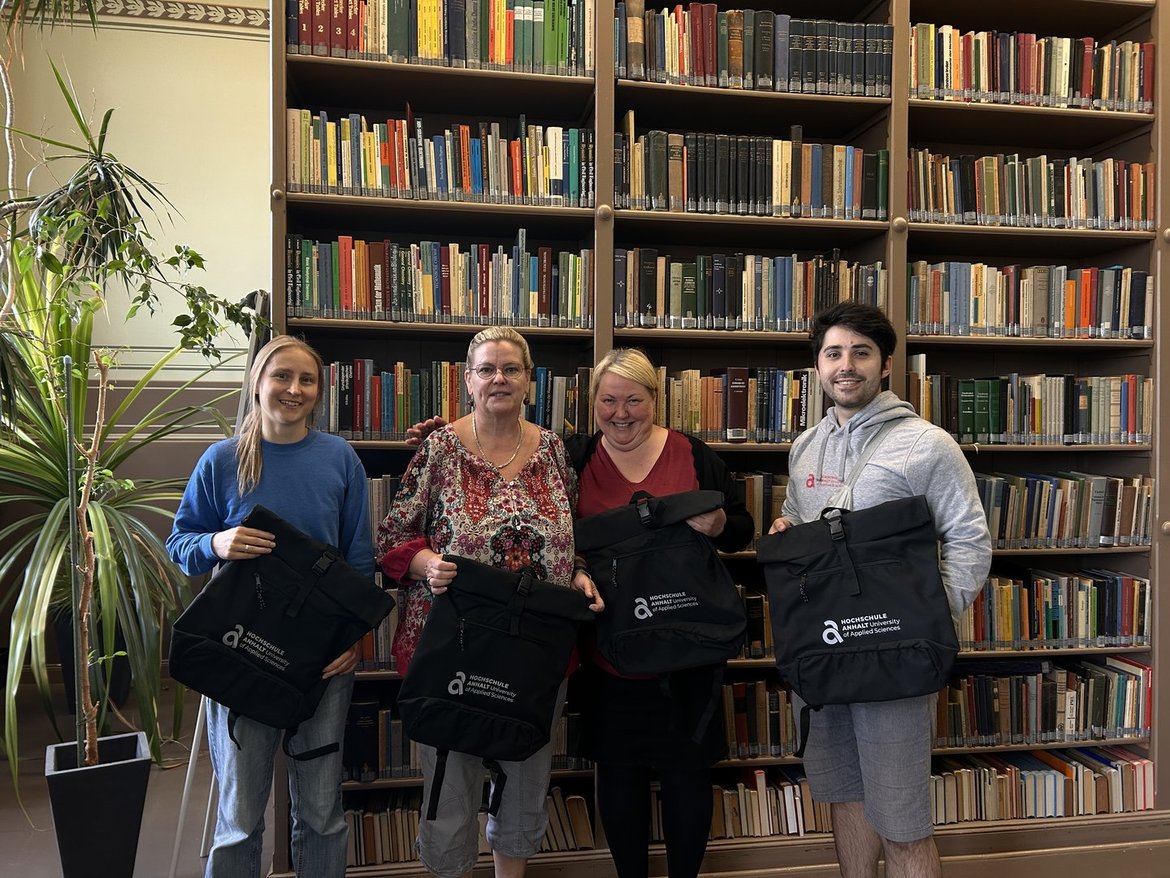 Vier Personen stehen lächelnd in einer Bibliothek vor einem Regal voller Bücher. Jede hält einen schwarzen Rucksack mit dem Logo der Deutschen Zentralbücherei für Blinde.