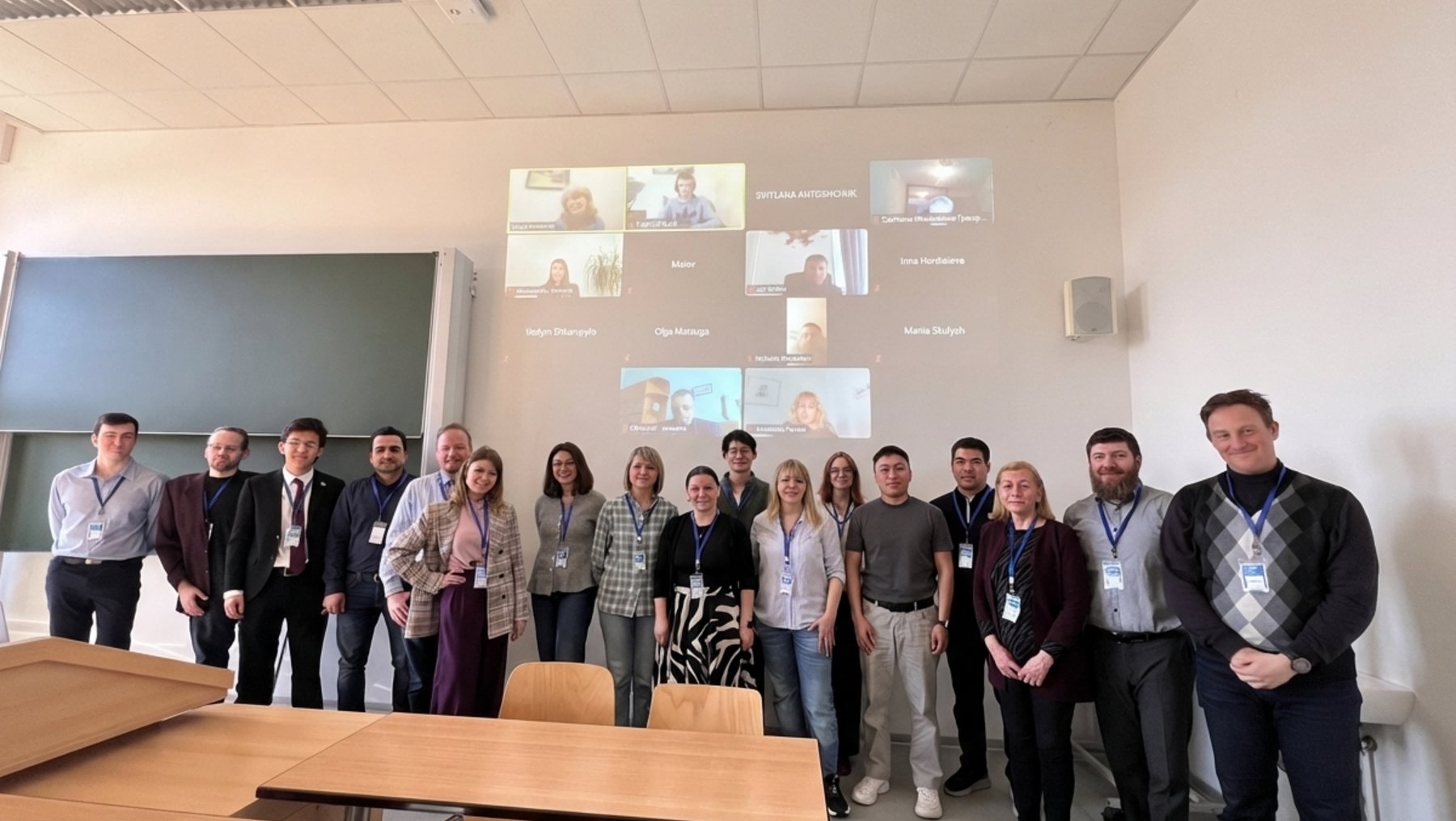 Gruppe von 16 Personen steht lächelnd in einem hellen Seminarraum, davon einige mit Namensschildern. Im Hintergrund sind auf einem Bildschirm vier Videochats mit weiteren Personen zu sehen.