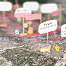 Miniaturmodell eines Campus mit markierten Bereichen wie Powerfeld, Re-Lab, Re-Hub, Mobi-Hub und weiteren Stationen, verbunden durch farbige Linien auf einer Landschaftsfläche.