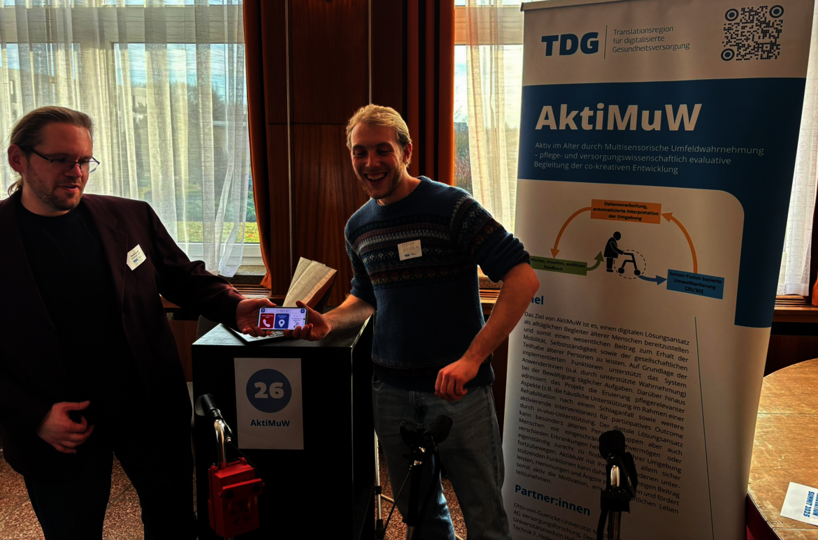Zwei Männer stehen an einem Infostand mit dem Namen AktiMuW, einem Projekt zur Mobilitätsförderung. Der Stand zeigt eine Person mit Rollstuhl und Textinfos. Einer hält ein Tablet, beide lächeln freundlich.