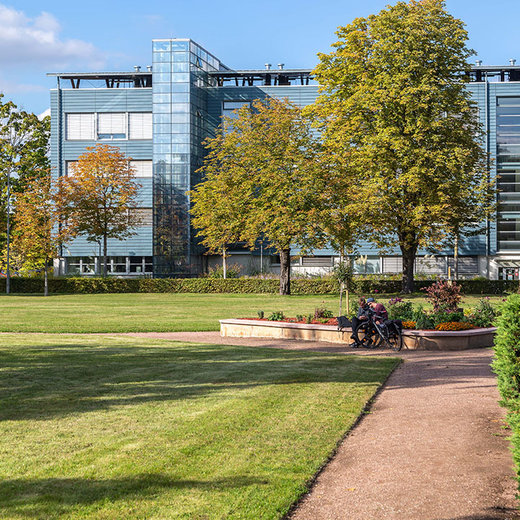 A modern office building with glass windows and a blue facade stands in the background. In the foreground, there is a well-maintained grassy area with a paved walkway. Two people are sitting on a bench surrounded by greenery and flowers. The scene is peaceful and inviting.