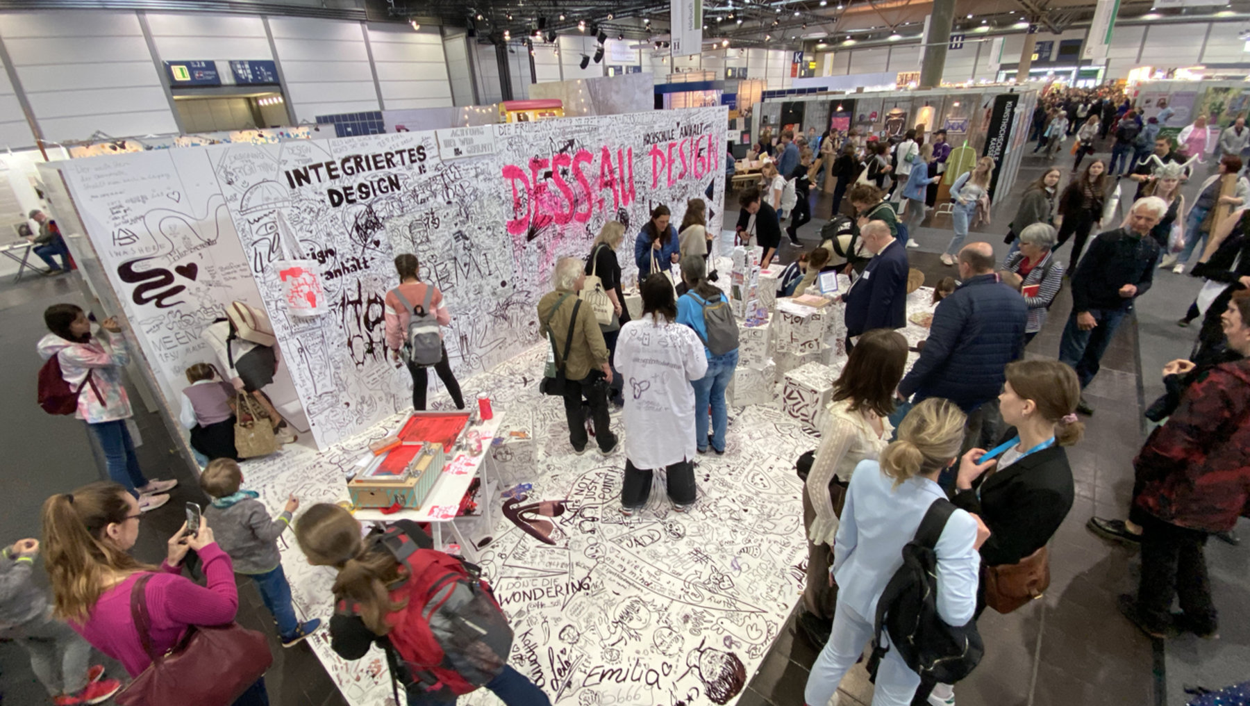 Große Menschenmenge vor einer mit Graffiti und Schriftzügen bedeckten Wand auf einer Messe. Die Beschriftung „Integriertes Design“ und „Dessau Design“ ist deutlich zu erkennen. Besucher:innen unterhalten sich, fotografieren oder schreiben selbst auf die Wand.