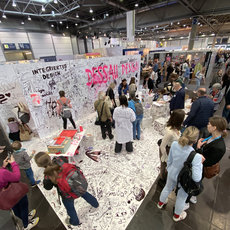 Große Menschenmenge vor einer mit Graffiti und Schriftzügen bedeckten Wand auf einer Messe. Die Beschriftung „Integriertes Design“ und „Dessau Design“ ist deutlich zu erkennen. Besucher:innen unterhalten sich, fotografieren oder schreiben selbst auf die Wand.