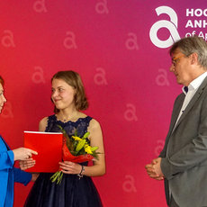 Eine junge Frau in dunklem Kleid mit Blumen erhält von einer Frau in Blau einen roten Ordner. Ein Mann in Anzug steht daneben, alle drei lächeln bei einer feierlichen Veranstaltung der Hochschule Anhalt.