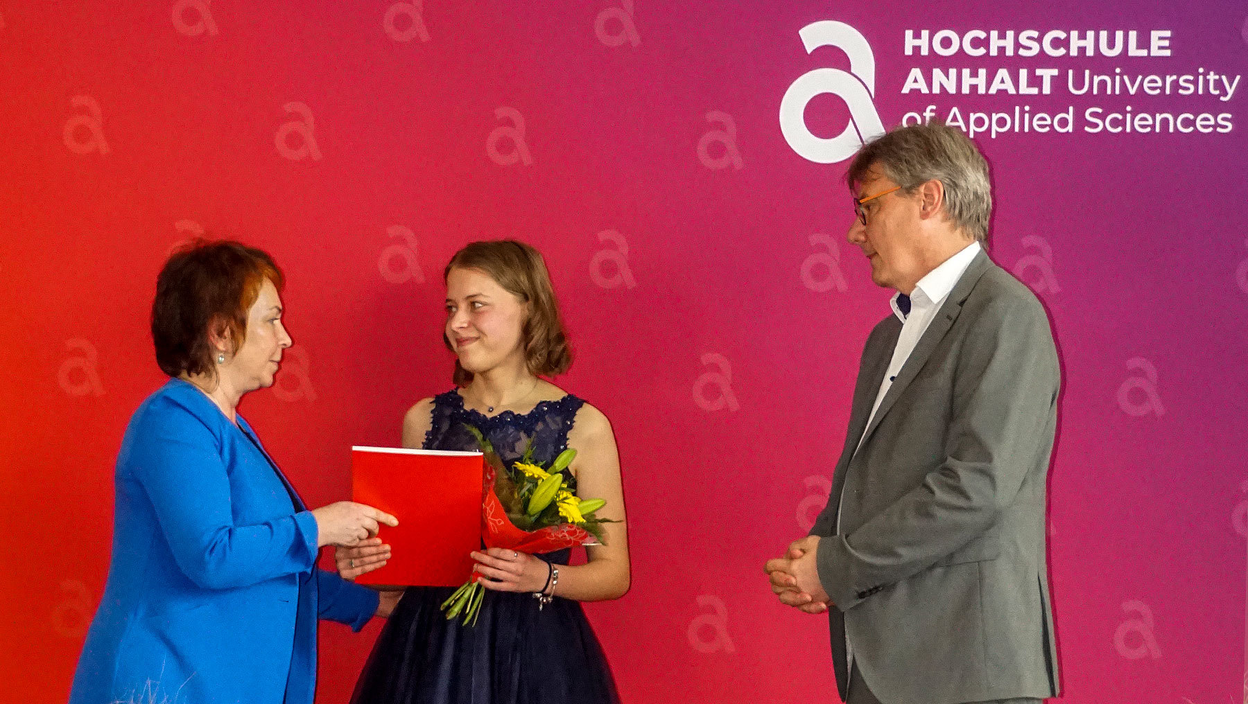 Eine junge Frau in dunklem Kleid mit Blumen erhält von einer Frau in Blau einen roten Ordner. Ein Mann in Anzug steht daneben, alle drei lächeln bei einer feierlichen Veranstaltung der Hochschule Anhalt.
