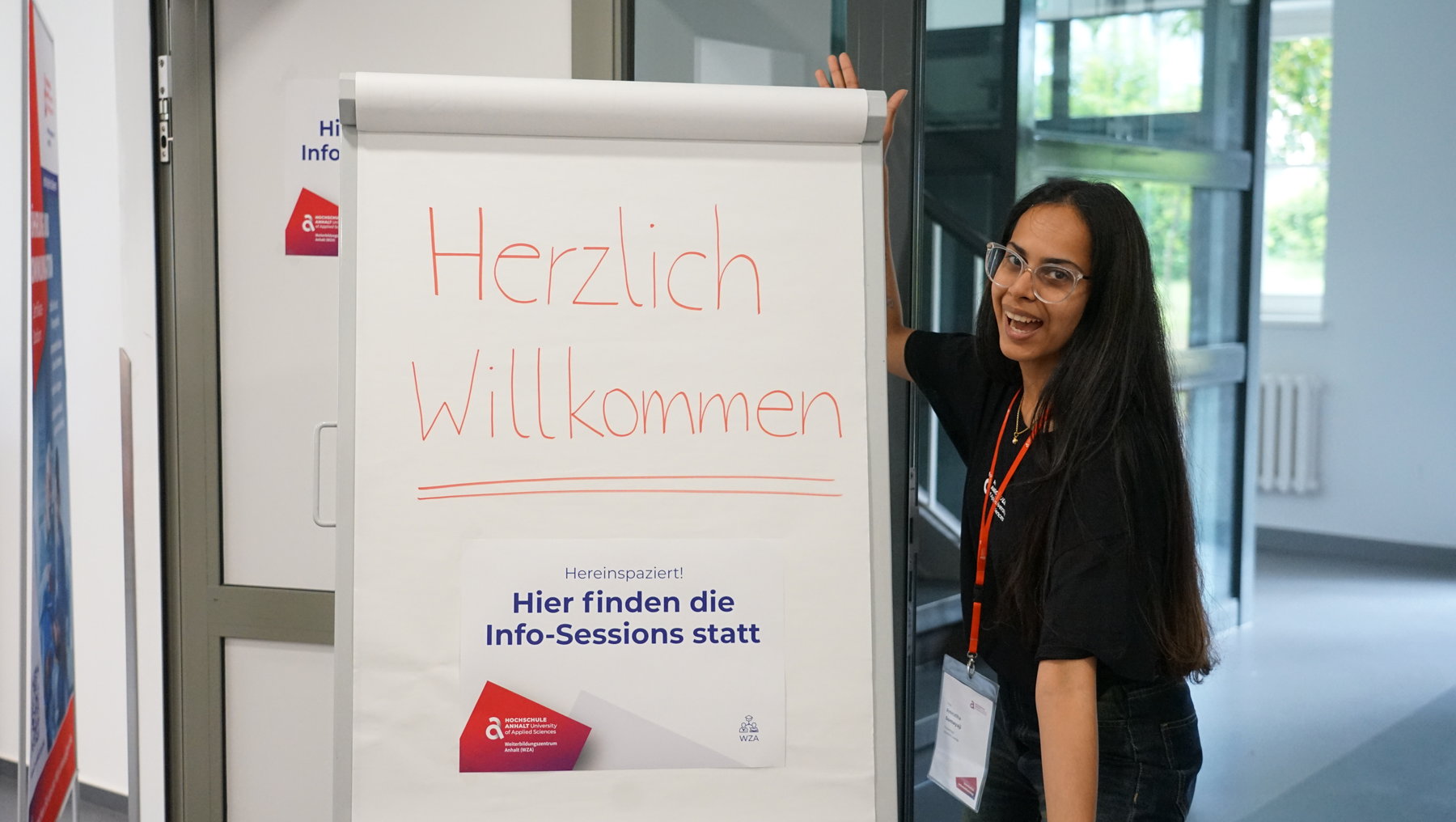 Eine lächelnde Person hält ein Plakat mit der Aufschrift „Herzlich Willkommen“ und darunter „Hier finden die Info-Sessions statt“.