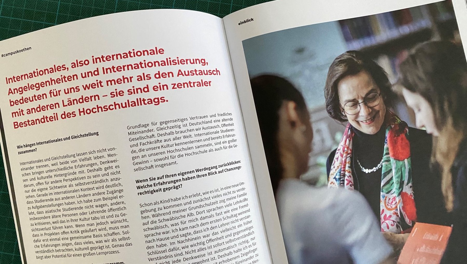 Eine lächelnde Frau mit Brille und rotem Pullover hält ein Schild mit der Aufschrift „Hochschultag“. Sie steht vor einem Hintergrund mit anderen Menschen, die ebenfalls an einer Veranstaltung teilnehmen. Der Artikel daneben thematisiert internationale Austauschprogramme und die Bedeutung von globaler Vernetzung im Hochschulalltag.