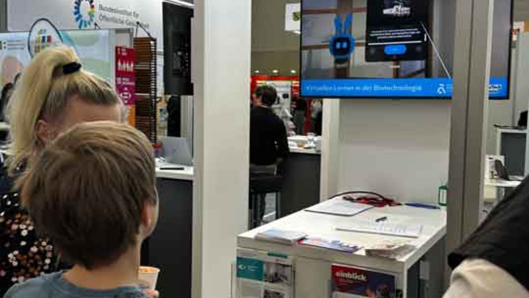 Ein Kind und eine erwachsene Person betrachten auf einer Messe einen Infostand zur Biotechnologie. Auf einem Tisch liegen Informationsmaterialien und Broschüren, während im Hintergrund ein Bildschirm mit einem Roboterarm und dem Thema „Lernen in der Biotechnologie“ zu sehen ist.