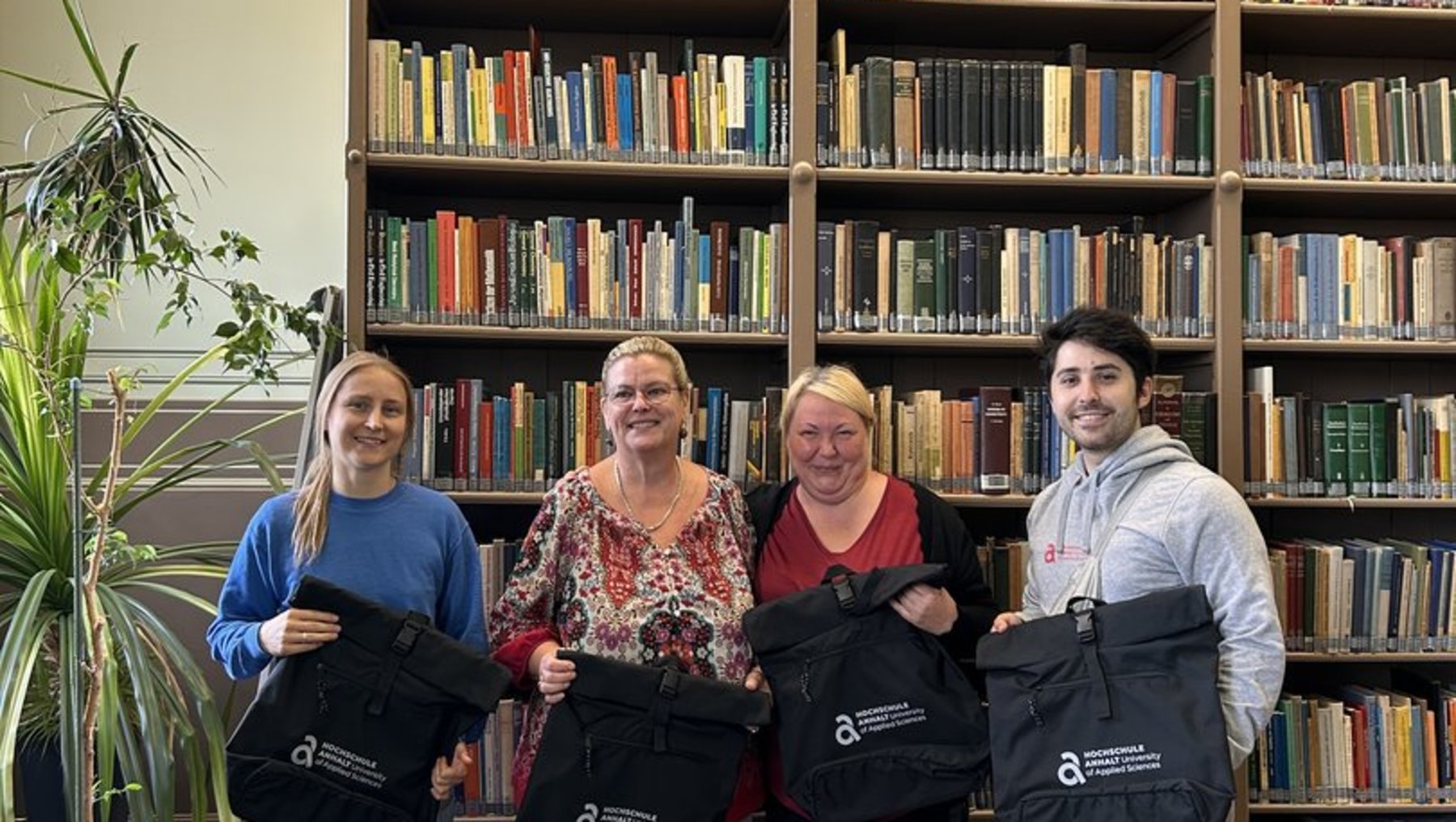 Vier Personen stehen vor einem Regal mit vielen bunten Büchern und halten jeweils eine schwarze Tasche mit einem Logo und Aufdruck. Sie lächeln und posieren gemeinsam für ein Foto.