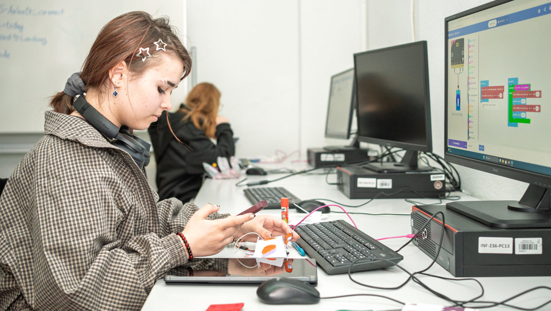 Eine Person baut am Schreibtisch ein kleines Elektronikprojekt mit Kabeln, Sensoren und einer Platine auf, während im Hintergrund ein Bildschirm mit einer Programmieroberfläche zu sehen ist.