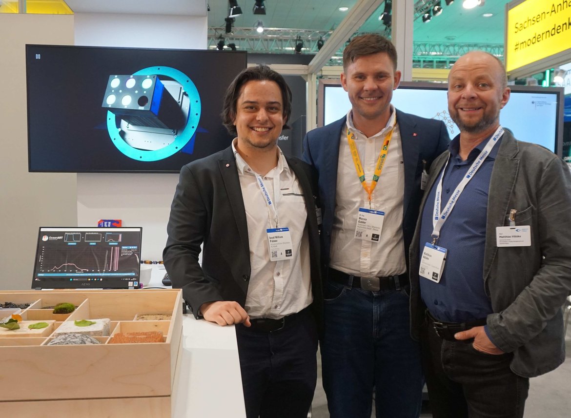 Drei Männer stehen lächelnd auf einer Messe. Hinter ihnen ist ein großer Bildschirm mit einem blauen, schematischen Rad oder Kreis zu sehen. Vor ihnen liegt ein Tisch mit verschiedenen kleinen Elektronikbauteilen und einem Laptop.
