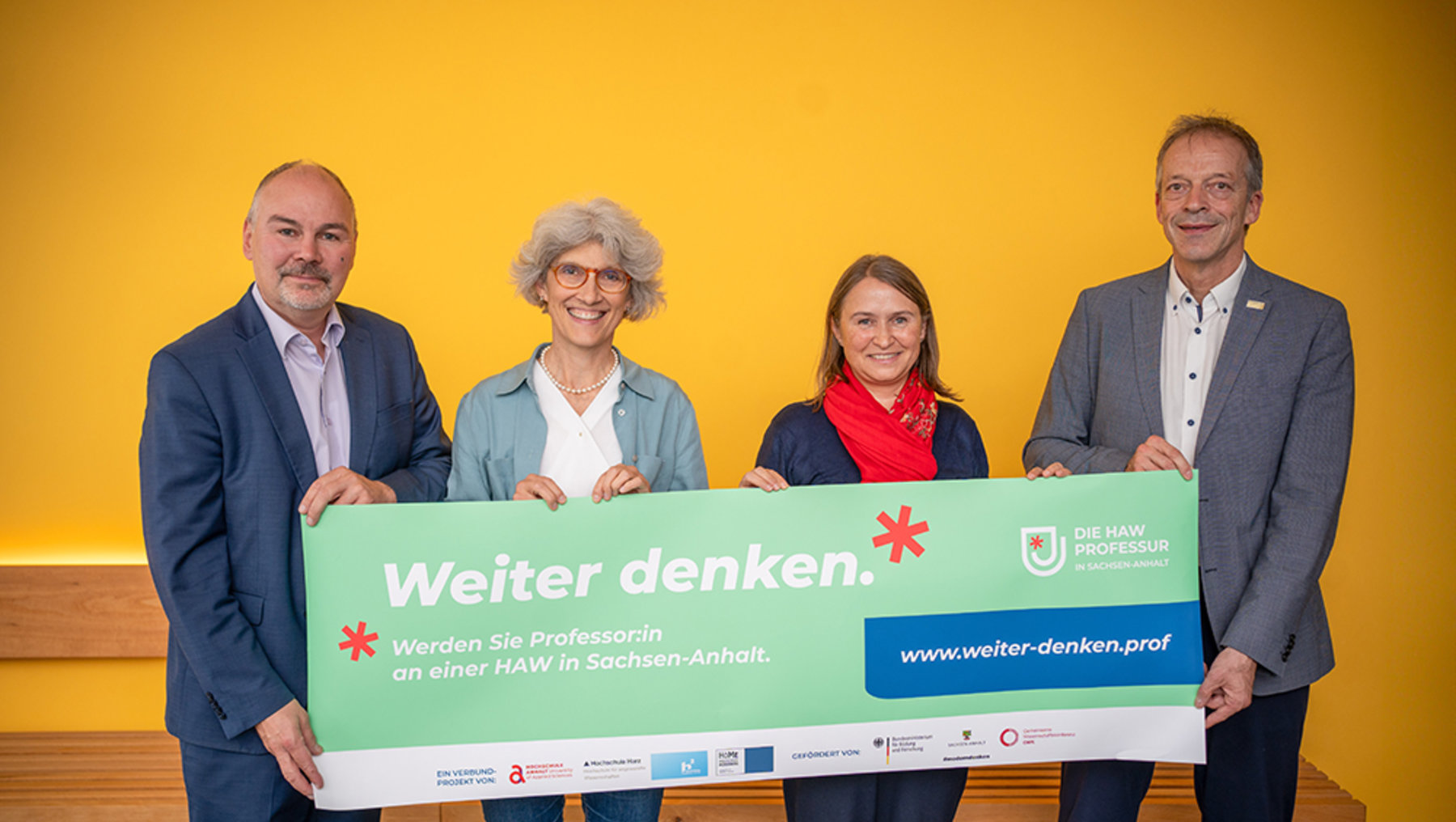 Vier Personen halten gemeinsam ein großes grünes Plakat mit der Aufschrift „Weiter denken.“ Darunter steht: „Werden Sie Professorin an einer HAW in Sachsen-Anhalt.“ Die Website wird mit „www.weiter-denken.prof“ angegeben.