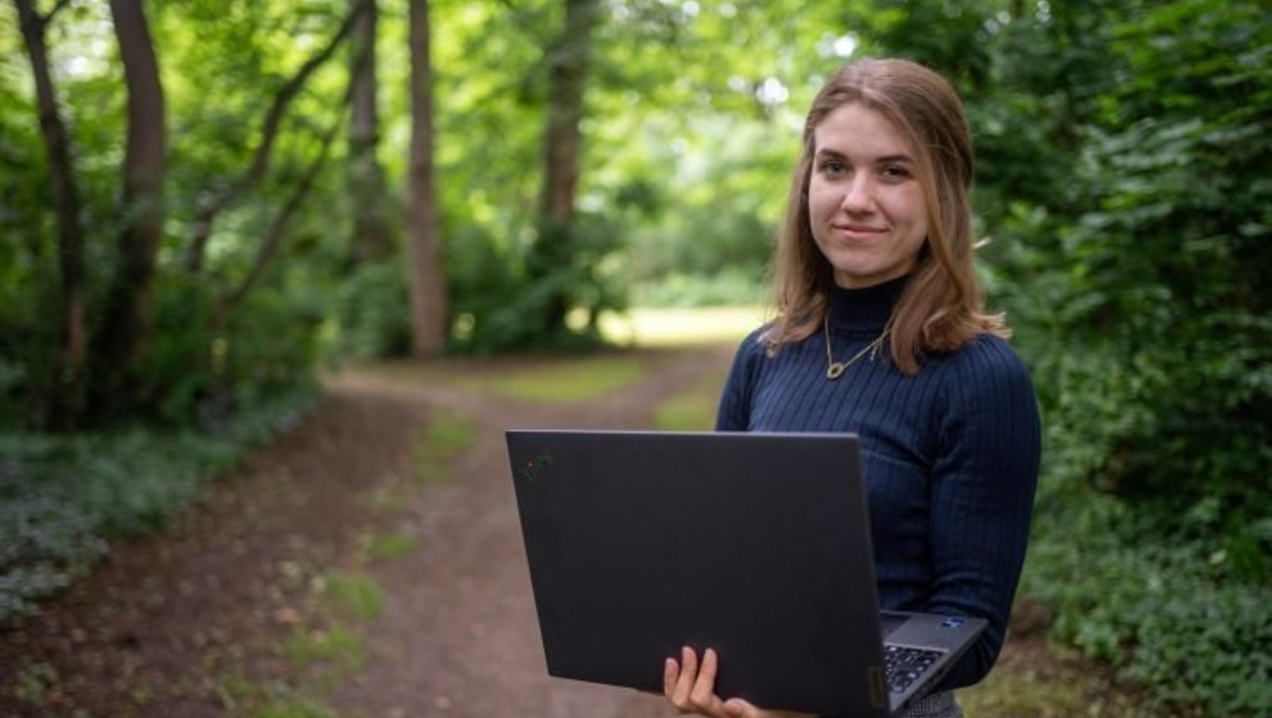 A woman stands on a forest path holding a black laptop. She is smiling and wearing a blue sweater and gray skirt. The background features green trees and foliage, creating a serene outdoor setting.