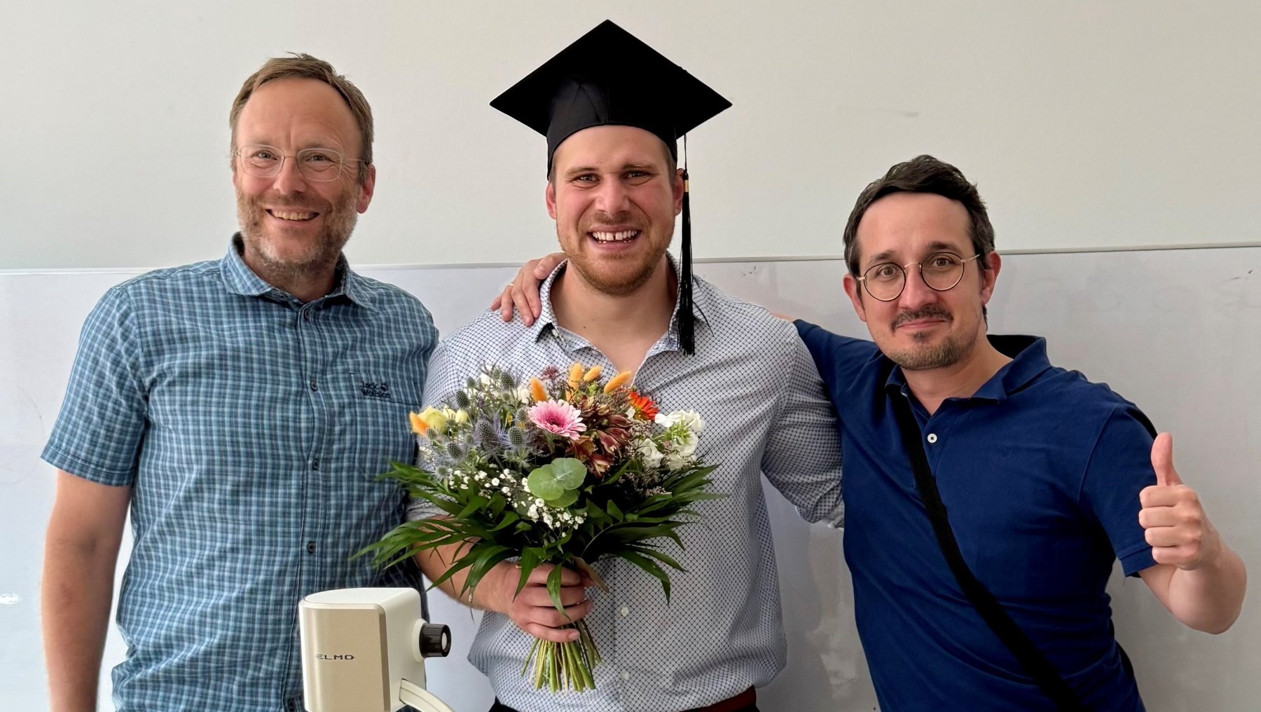 Drei Männer stehen lächelnd beieinander: Der Mann in der Mitte trägt eine schwarze Graduationsmütze und hält einen Strauß Blumen. Links und rechts stehen zwei weitere Männer mit Daumen hoch, die ihn freudig umarmen.