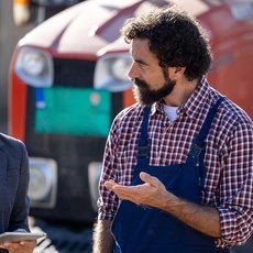 Eine Frau in Business-Kleidung und ein Mann mit kariertem Hemd und Arbeitshose unterhalten sich auf einer Baustelle, während im Hintergrund ein roter Bagger zu sehen ist.