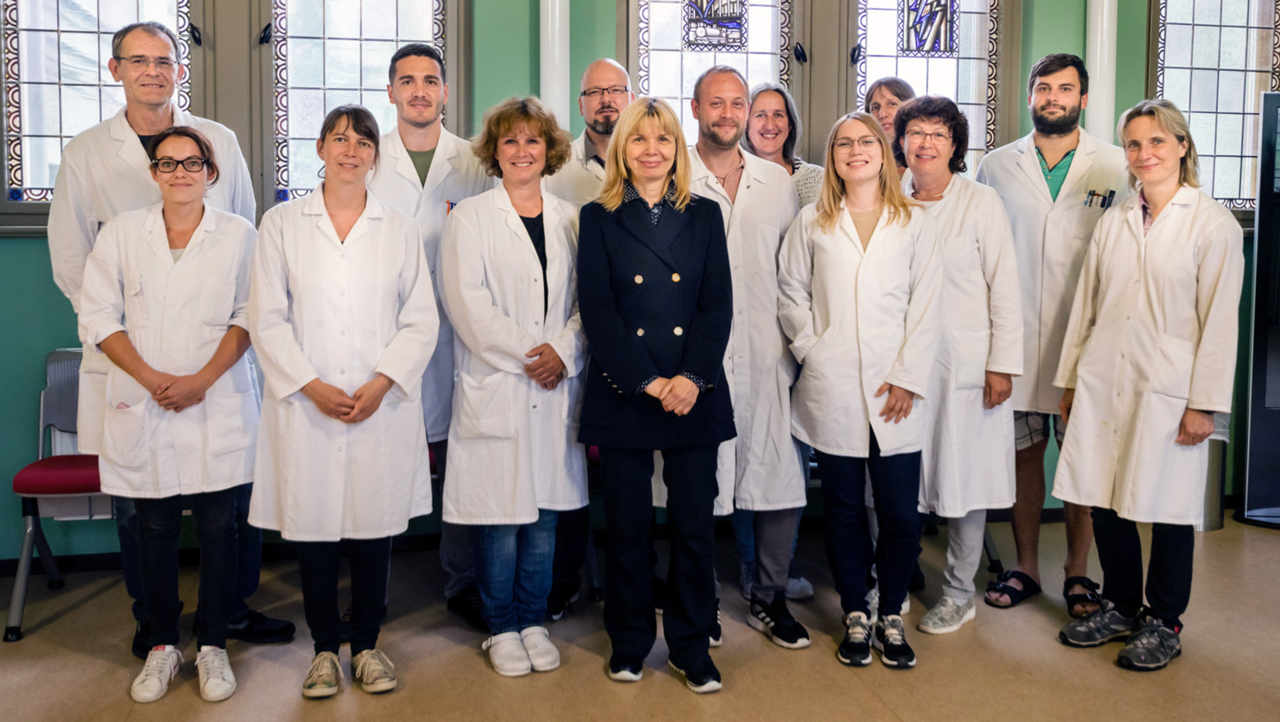 A group of fifteen medical professionals dressed in lab coats poses together indoors. One individual sits in the front row wearing casual clothes. Everyone appears ready, likely for a team or group photo.