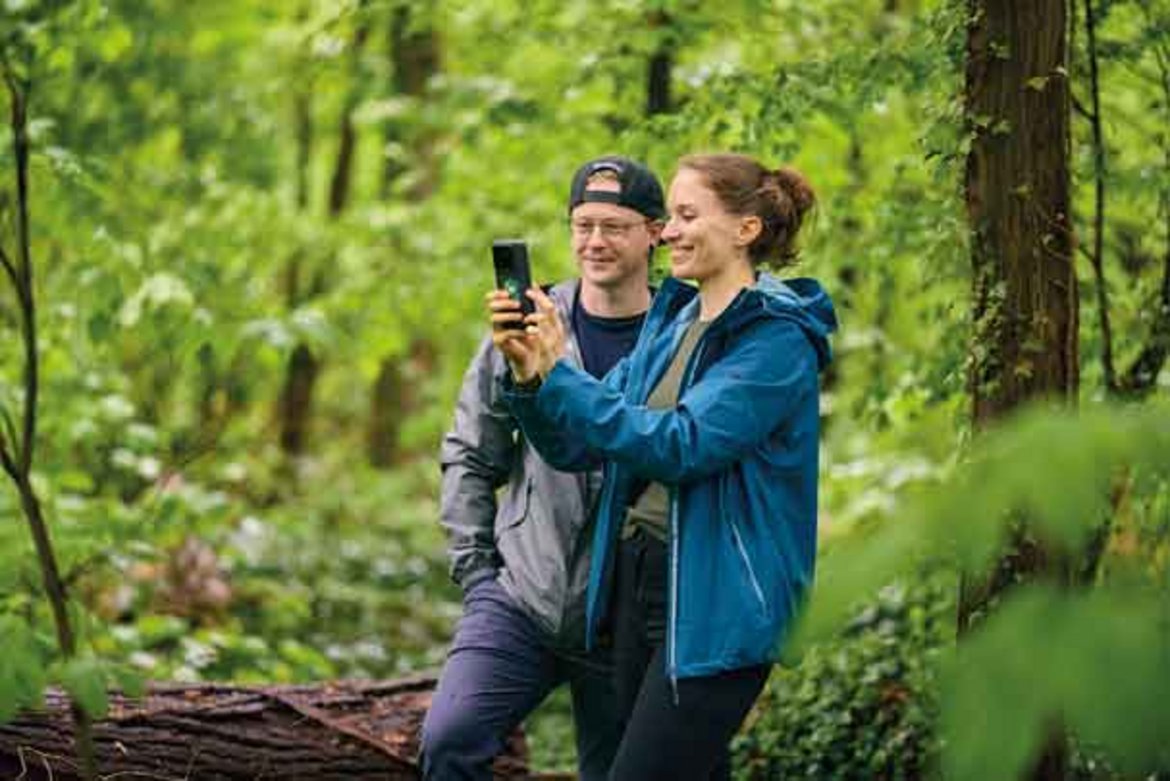 Zwei Personen im Wald, die gemeinsam ein Selfie mit dem Handy machen. Die Umgebung ist grün und dicht bewachsen mit Bäumen und Farnen, ein sonniger Waldweg ist im Hintergrund zu sehen.