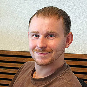 Lächelnder Mann mit kurzem Bart und kurzem Haar, der ein braunes T-Shirt trägt und freundlich in die Kamera schaut.