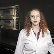 Eine Person mit lockigen, hellbraunen Haaren und Brille steht lächelnd in einer Küche. Sie trägt einen weißen Laborkittel und steht vor einem doppelten Backofen.