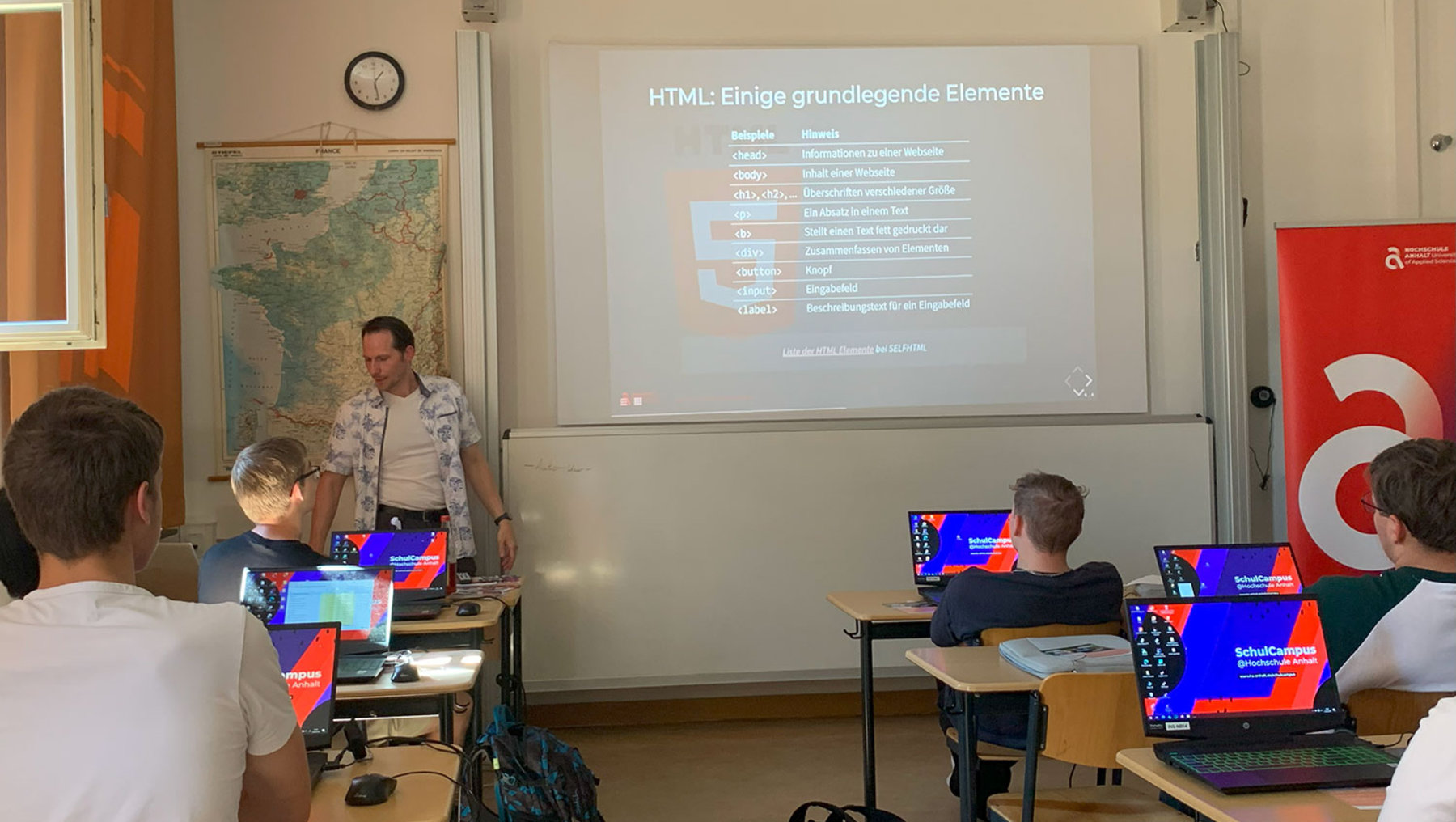 Ein Dozent steht vor einer Klasse und erklärt an einer Tafel oder Leinwand grundlegende HTML-Elemente wie Überschriften, Absätze und Listen. Die Schüler arbeiten an Laptops mit einem farbigen Bildschirmhintergrund.