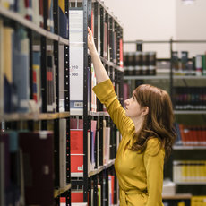 A person in a yellow jacket reaches upward on tiptoes to access a high shelf in a well-stocked library or bookstore, surrounded by tall bookshelves filled with colorful books.