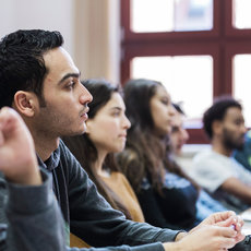 A group of students sitting in a classroom, attentively listening and participating in a discussion. The students are seated in a row, with one student raising his hand. The setting appears to be indoors with natural light coming through the windows.