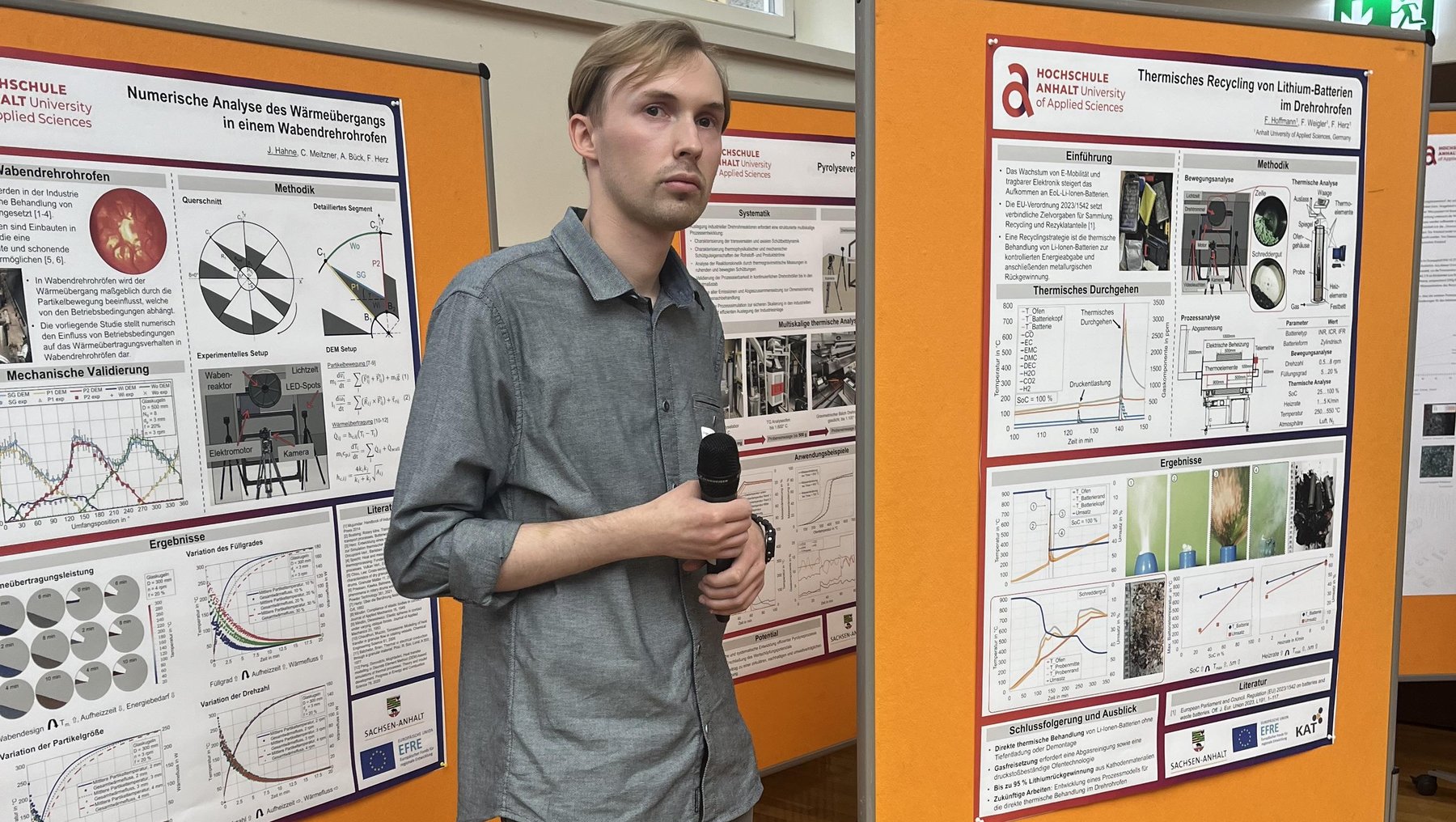 Ein junger Mann steht vor zwei orangefarbenen Posterwänden mit wissenschaftlichen Präsentationen. Er hält ein Mikrofon und präsentiert seine Ergebnisse zu einem Thema mit Diagrammen, Grafiken und Texten, darunter Titel wie „Superresolution“ und „Analyse der Messdaten“.