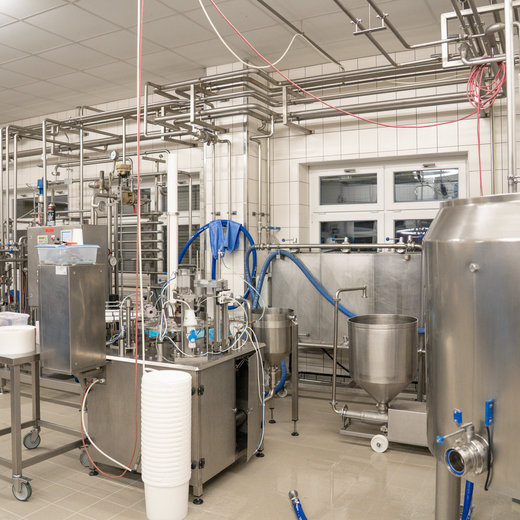 Moderne Brauerei oder Lebensmittelproduktionsanlage mit Edelstahltanks, Rohrleitungen und verschiedenen Maschinen zur Flüssigkeitsverarbeitung. Saubere, helle Arbeitsumgebung mit Hygienestandards.