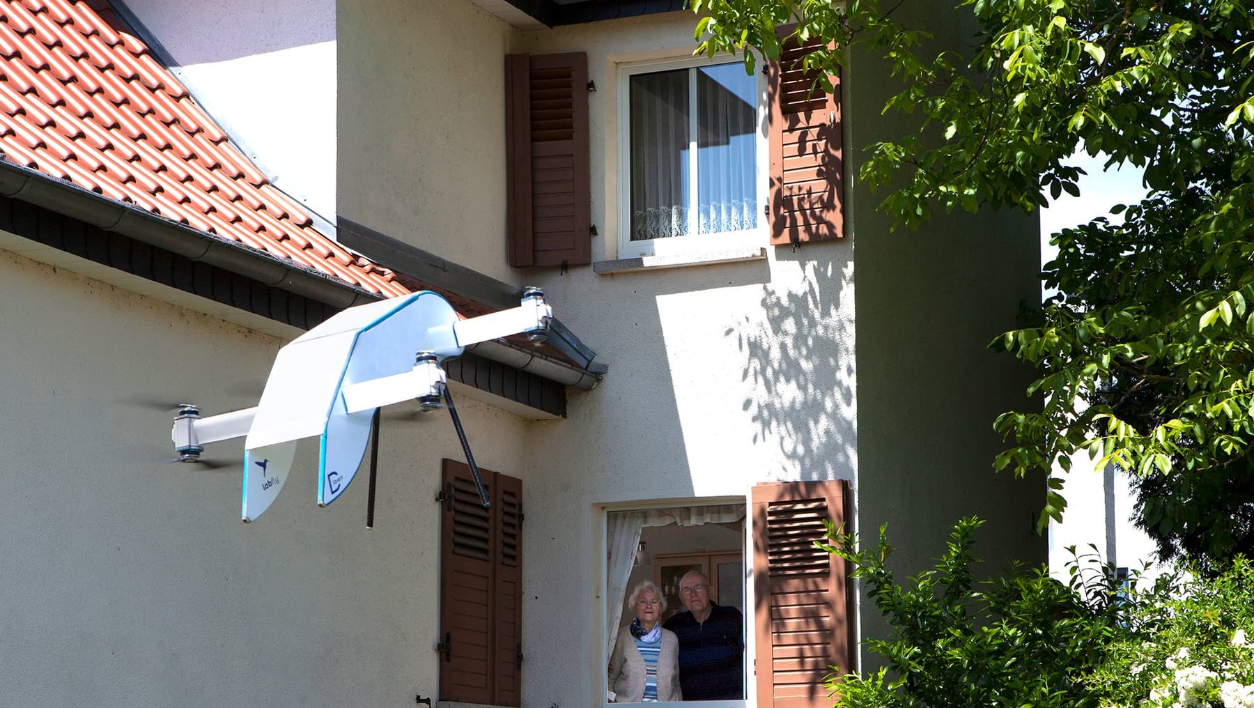 Zwei Personen stehen in einem Fenster des Erdgeschosses eines Hauses. Sie lächeln und schauen in Richtung eines uttryklinischen Satellitenschüssels, der an der Hauswand befestigt ist. Die Szene wirkt freundlich und einladend.