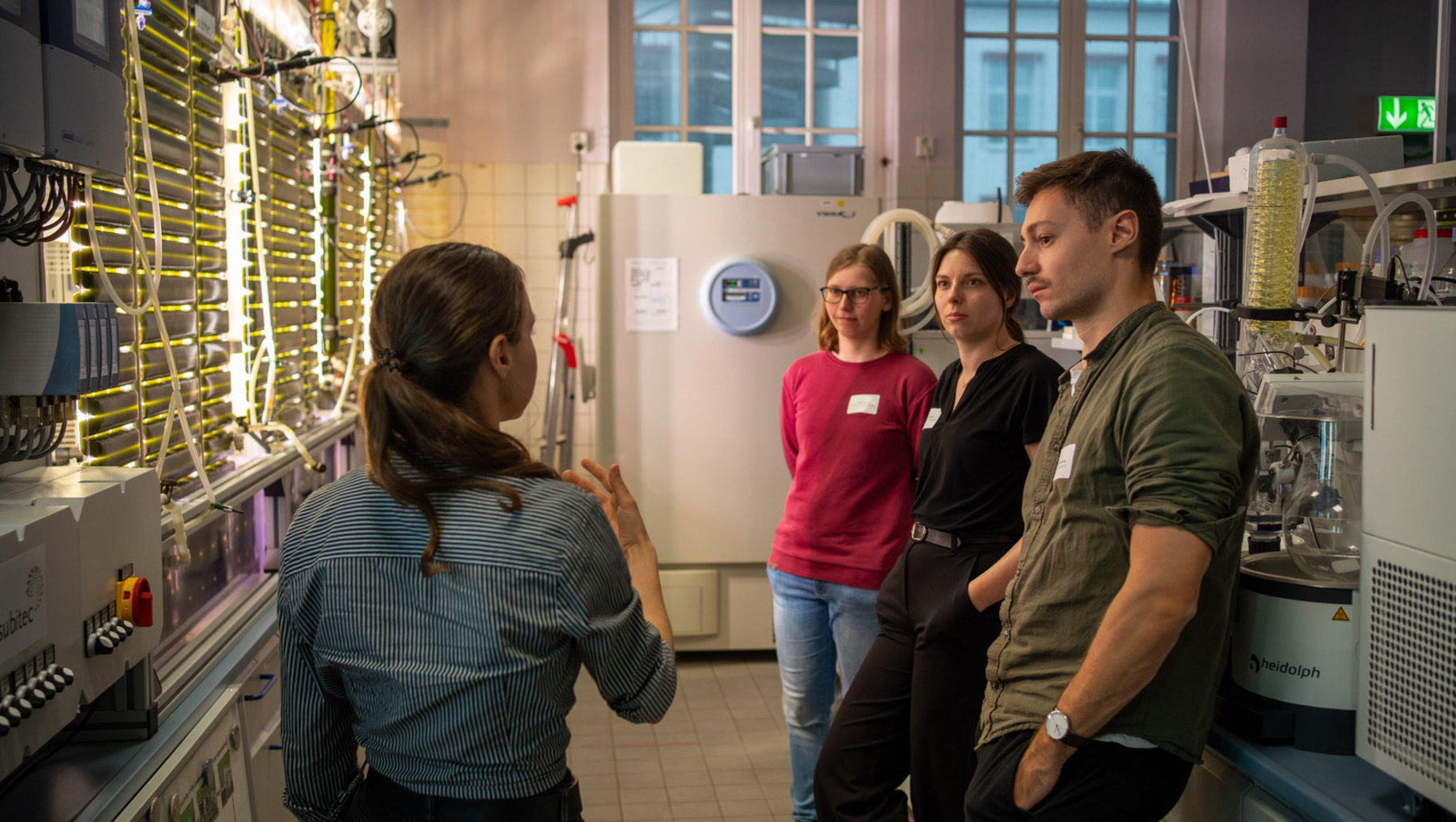 Eine Person erklärt in einem Labor vier Zuhörern mit Namensschildern eine wissenschaftliche Apparatur, während sie auf eine große Metallvorrichtung zeigt.