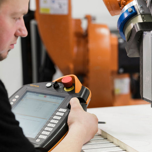 A man operates a handheld digital device near a robotic arm in a manufacturing setting. The device appears to be used for programming or monitoring the robotic arm, which is positioned over a work surface. The scene suggests precision engineering or automated manufacturing processes.
