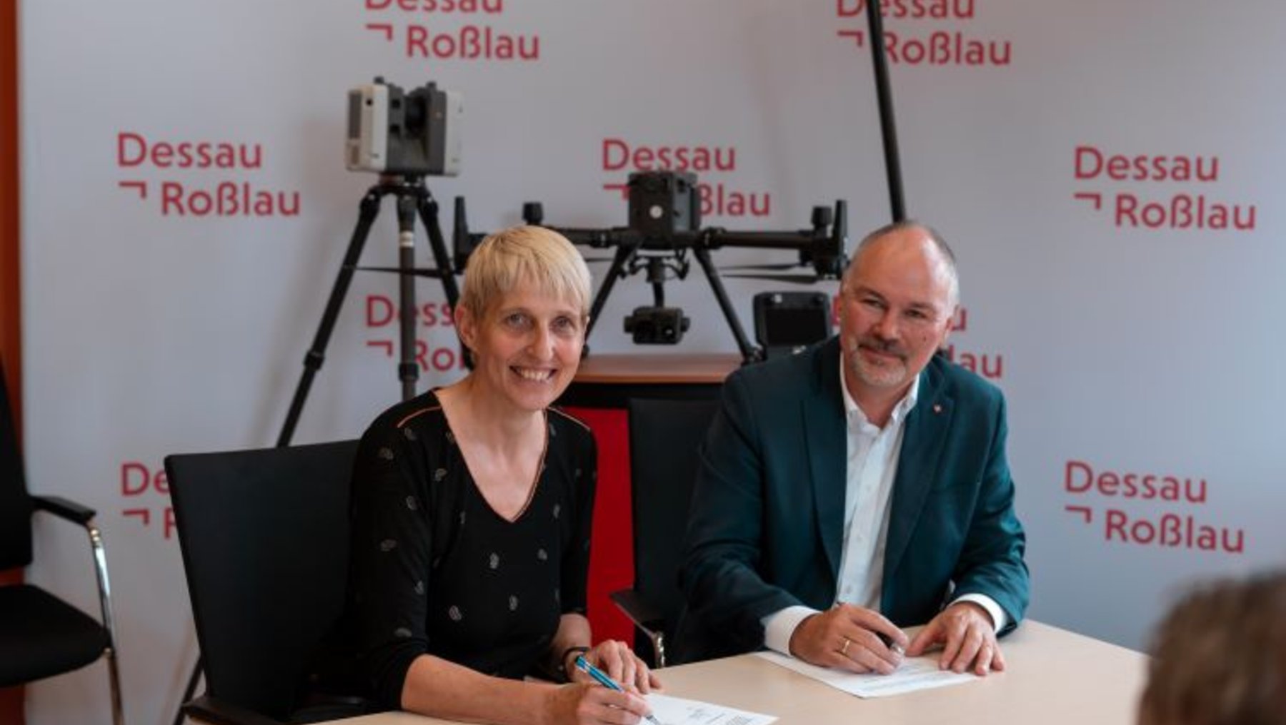 Zwei Personen sitzen an einem Tisch und unterschreiben ein Dokument. Im Hintergrund steht "Dessau Roßlau". Eine Kamera ist auf sie gerichtet. Die Szene wirkt offiziell, möglicherweise handelt es sich um eine Vertragsunterzeichnung oder eine ähnliche Zeremonie.