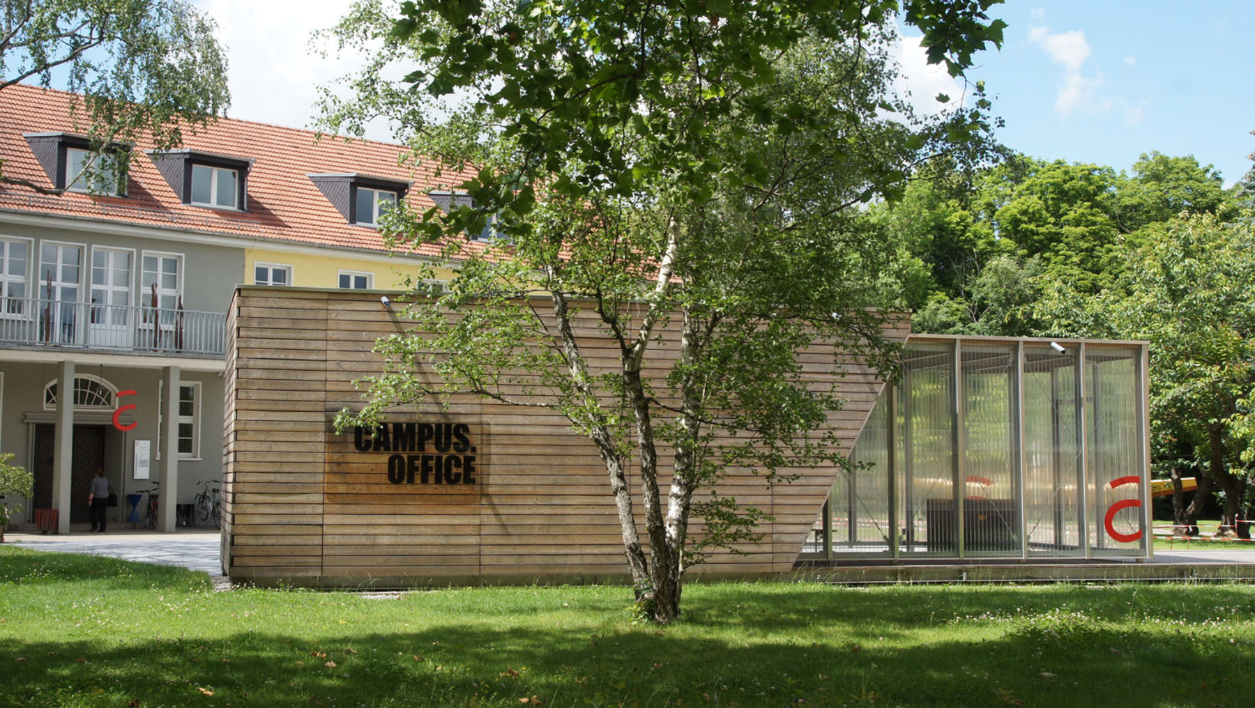 "Ein Campus-Bürogebäude mit einem modernen Anbau aus Holz und Glas. Im Hintergrund befindet sich ein älteres Gebäude mit einem roten Dach. Vor dem Gebäude steht ein Baum auf einem grünen Rasen."